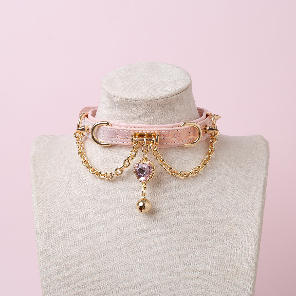 Pink leather choker featuring gold tone chains, D rings, spike studs, bell and pink and gold crystal heart charm. Displayed on a white mannequin bust against a pink background for a kawaii fashion aesthetic.