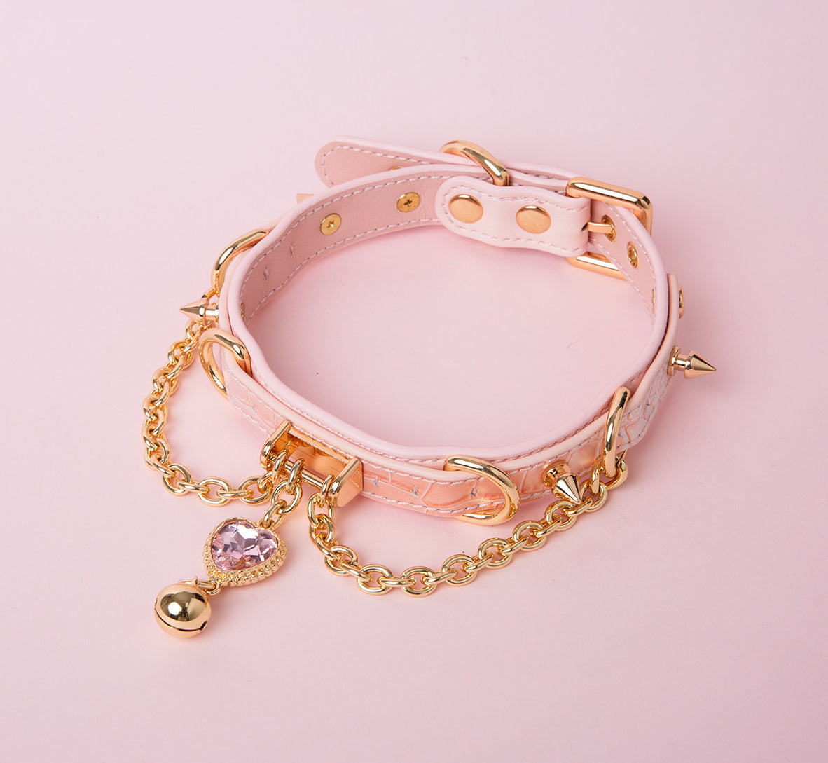 Pink leather choker featuring gold tone chains, D rings, spike studs, bell and pink and gold crystal heart charm. Displayed on a pink background for a Harajuku fashion aesthetic.