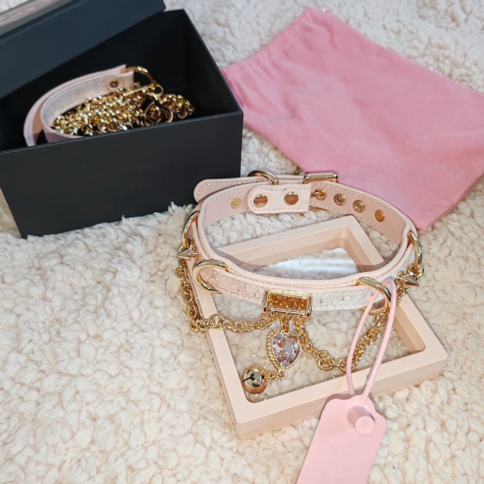 Pink leather choker and leash set featuring gold tone chains, D rings, spike studs, bell and pink and gold crystal heart charm, with detachable chain leash and leather hand strap. Displayed in a black and pink storage box for a casual lifestyle shot.