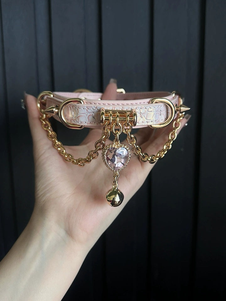 Pink leather choker featuring gold tone chains, D rings, spike studs, bell and pink and gold crystal heart charm. Held by a hand against a black wooden background for a pastel goth aesthetic.