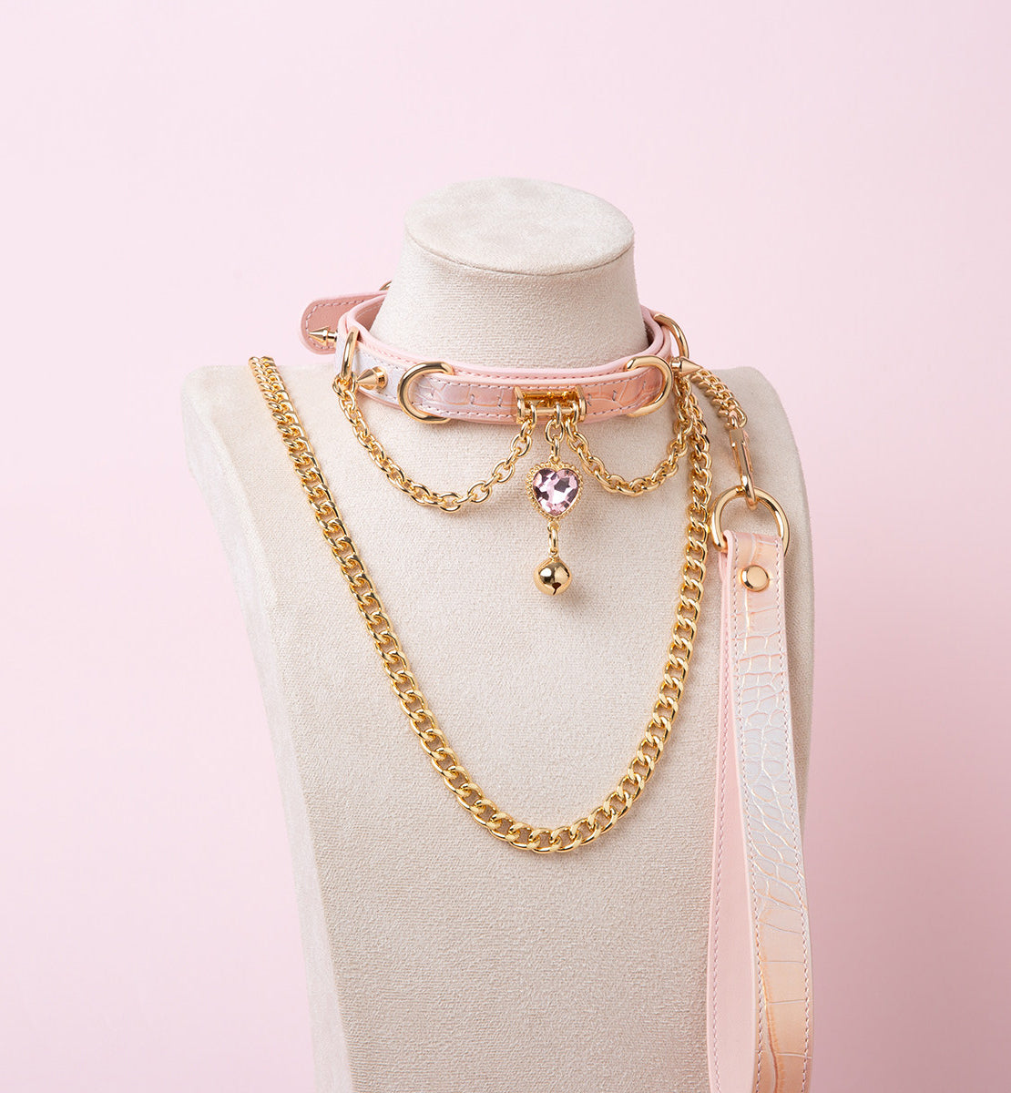 Pink leather choker and leash set featuring gold tone chains, D rings, spike studs, bell and pink and gold crystal heart charm, with detachable chain leash and leather hand strap. Displayed on a white mannequin bust against a pink background for a kawaii goth aesthetic.