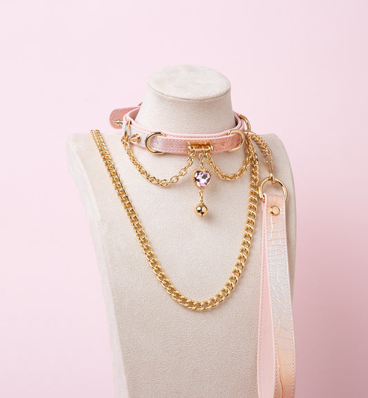 Pink leather choker and leash set featuring gold tone chains, D rings, spike studs, bell and pink and gold crystal heart charm, with detachable chain leash and leather hand strap. Displayed on a white mannequin bust against a pink background for a kawaii goth aesthetic.