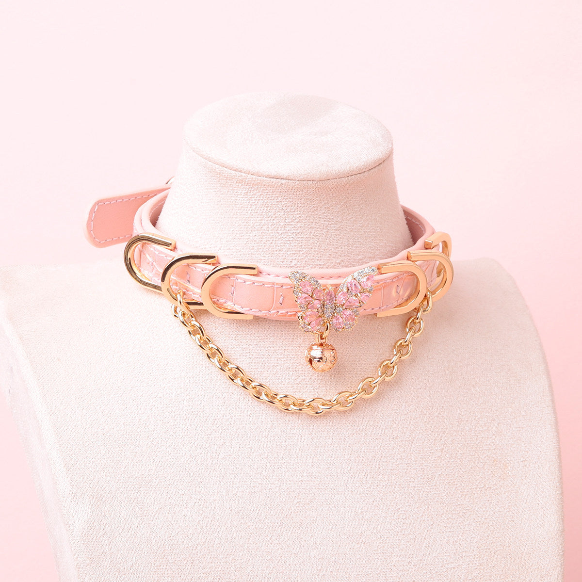 Pink leather choker featuring a gold and pink diamante butterfly charm, chains, D rings and bell. Displayed on a white mannequin bust against a pink background for a kawaii fashion aesthetic.