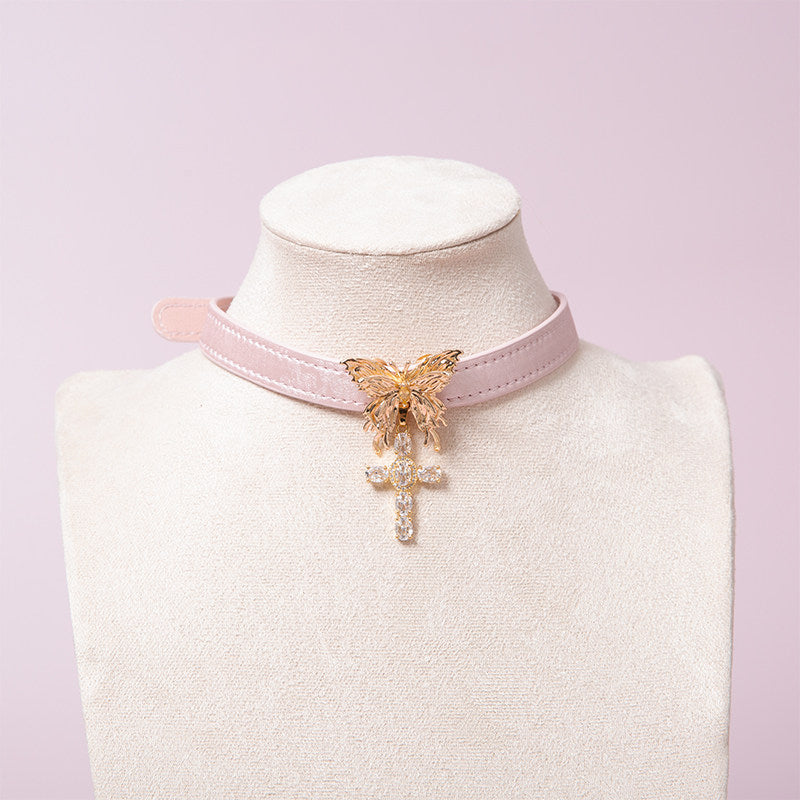 Pink leather choker featuring an intricate gold tone butterfly pendant and gold and clear diamante cross charm. Displayed on a white mannequin bust against a pink background for a Harajuku fashion aesthetic.