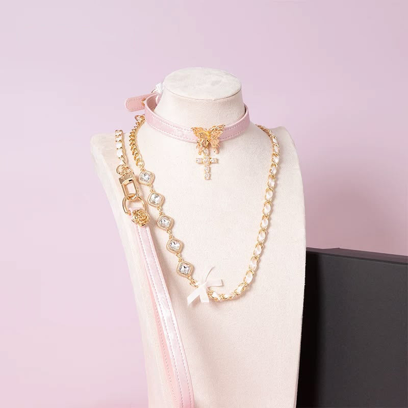 Pink leather choker featuring an intricate gold tone butterfly pendant and gold and clear diamante cross charm, with detachable gold and crystal chain leash and pink leather hand strap. Displayed on a white mannequin bust against a pink background for a pastel goth aesthetic.