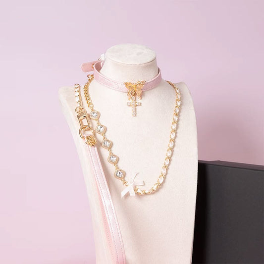Pink leather choker featuring an intricate gold tone butterfly pendant and gold and clear diamante cross charm, with detachable gold and crystal chain leash and pink leather hand strap. Displayed on a white mannequin bust against a pink background for a pastel goth aesthetic.