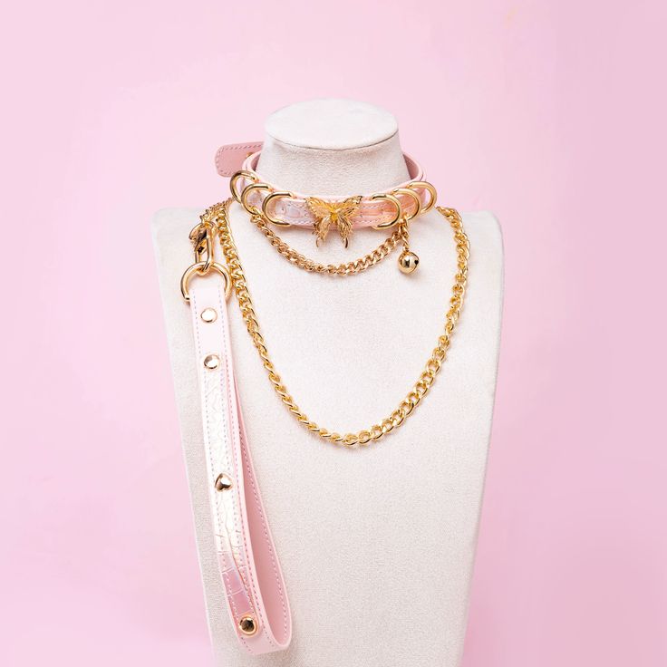 Pink leather choker and leash set featuring gold tone butterfly charm, D rings, chain and bell with detachable chain leash and leather hand strap. Displayed on a white mannequin bust against a pink background for a pastel goth aesthetic.