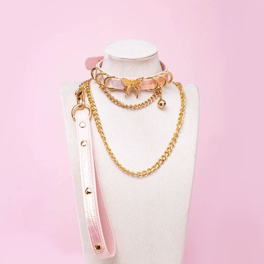 Pink leather choker and leash set featuring gold tone butterfly charm, D rings, chain and bell with detachable chain leash and leather hand strap. Displayed on a white mannequin bust against a pink background for a pastel goth aesthetic.