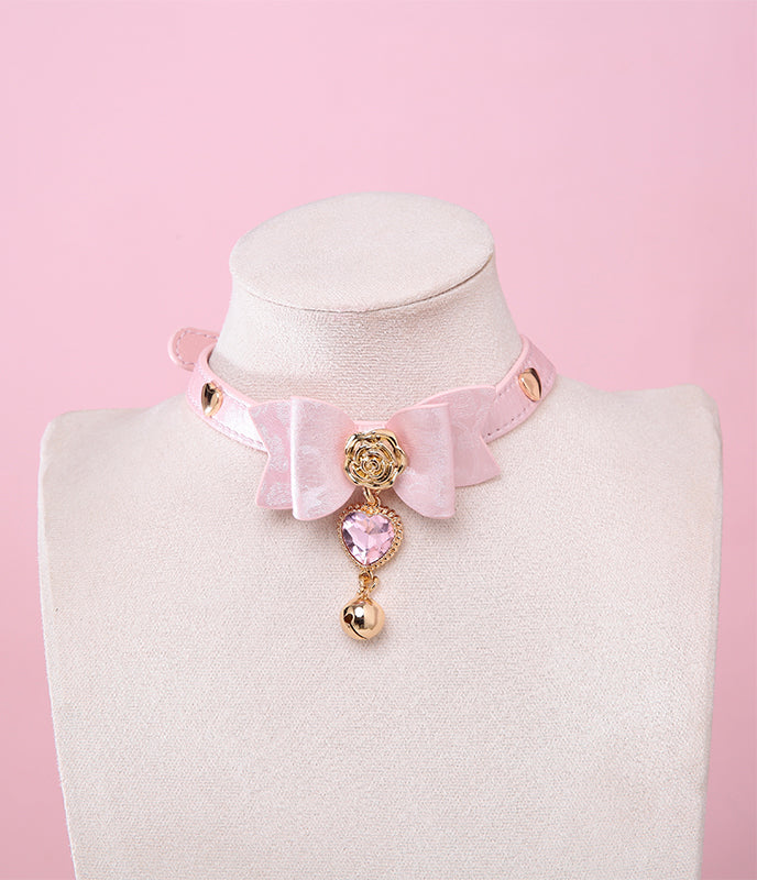 Pink leather choker featuring a central bow shaped design, gold tone rose and heart studs with detachable pink and gold crystal heart and bell charm. Displayed on a white mannequin bust against a pink background for a kawaii fashion aesthetic.