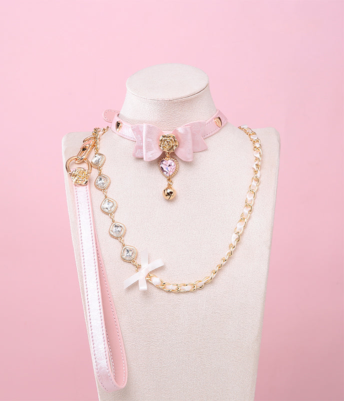 Pink leather choker and leash set featuring a central bow shaped design, gold tone rose and heart studs, pink and gold crystal heart and bell charm with detachable diamante, ribbon and chain leash and leather hand strap. Displayed on a white mannequin bust against a pink background for a pastel goth aesthetic.