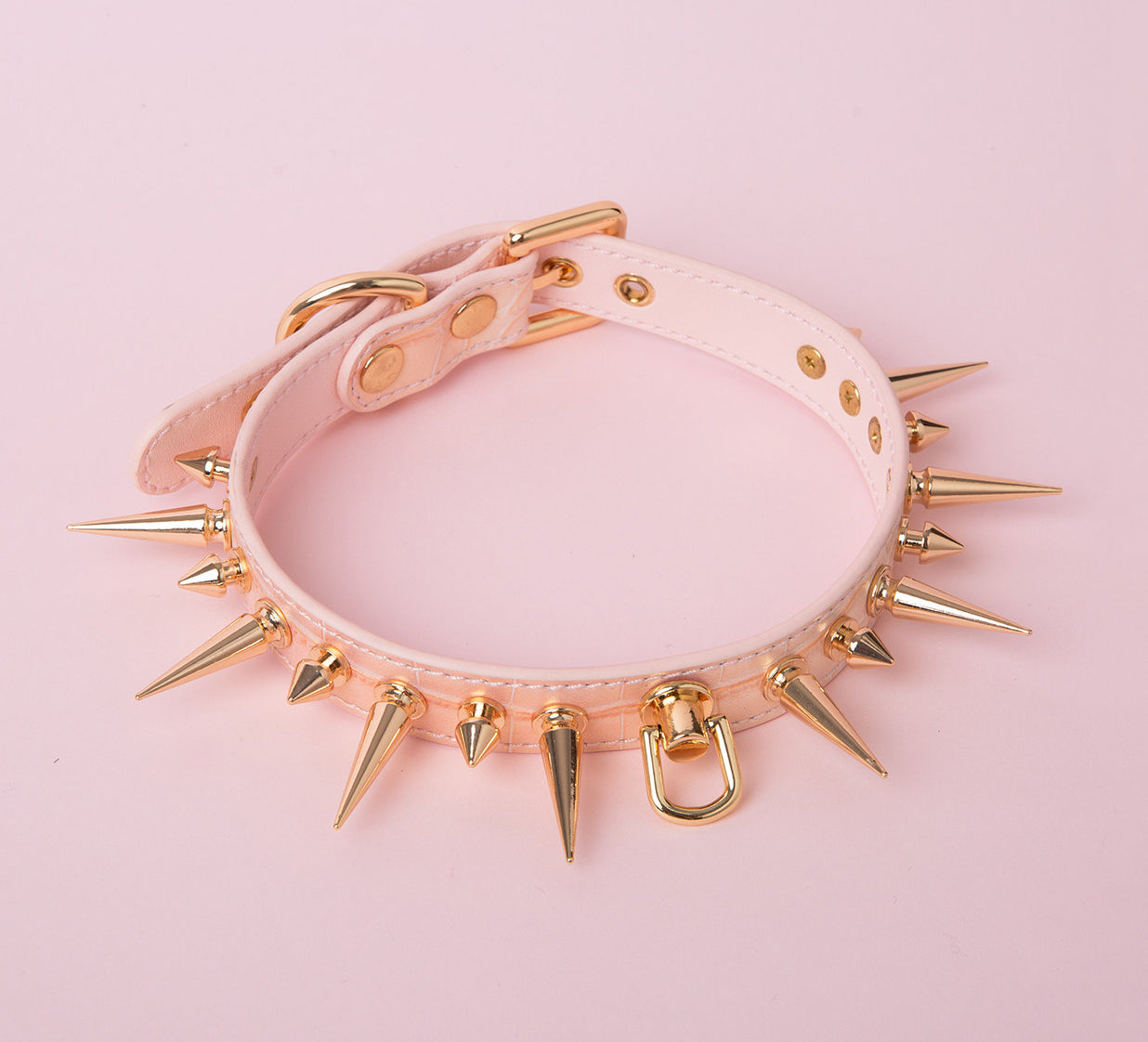 Pink faux crocodile effect leather choker featuring gold tone alternating length spike studs and central D ring attachment. Displayed on a pink background for a kawaii goth aesthetic.