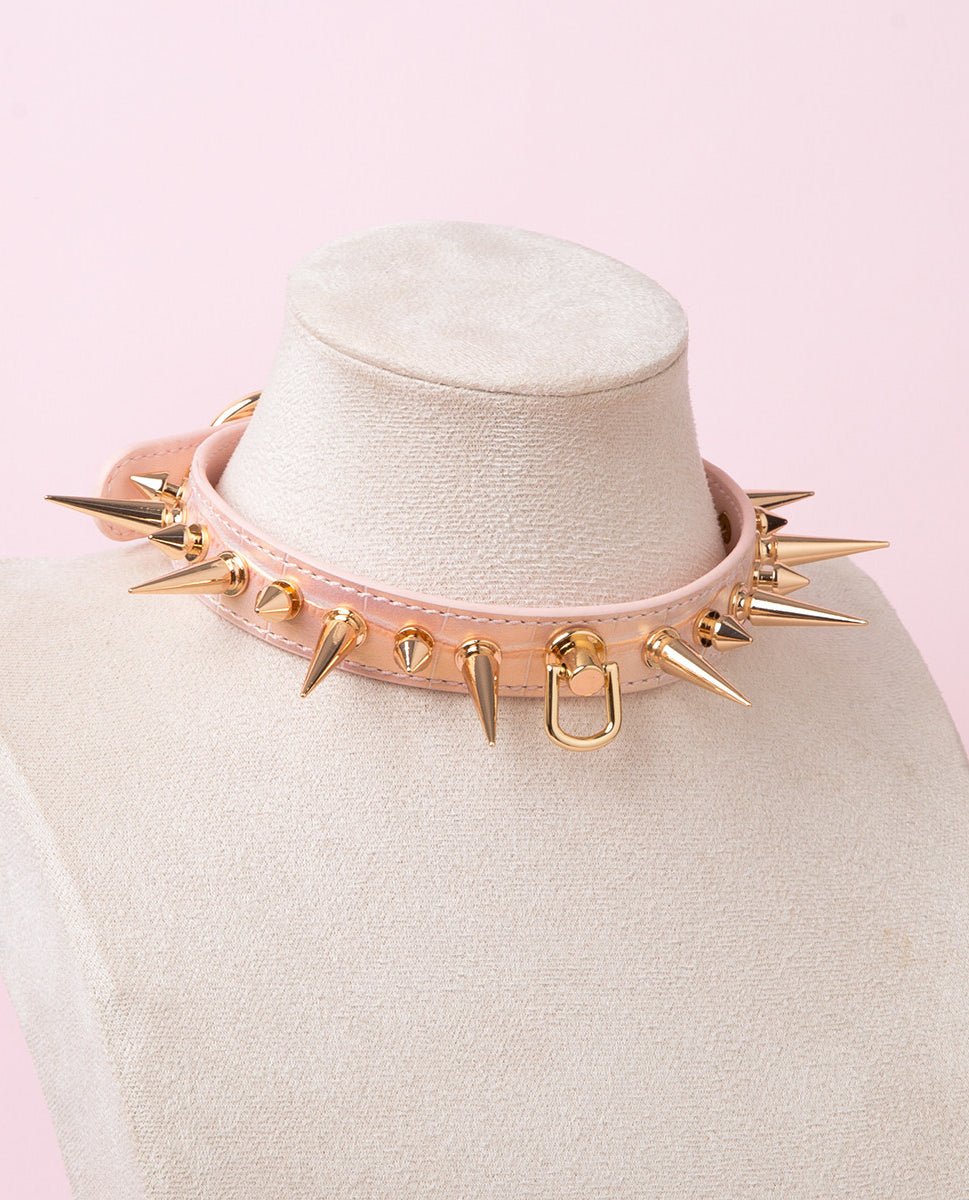 Pink faux crocodile effect leather choker featuring gold tone alternating length spike studs and central D ring attachment. Displayed on a white mannequin bust against a pink background for a kawaii goth aesthetic.