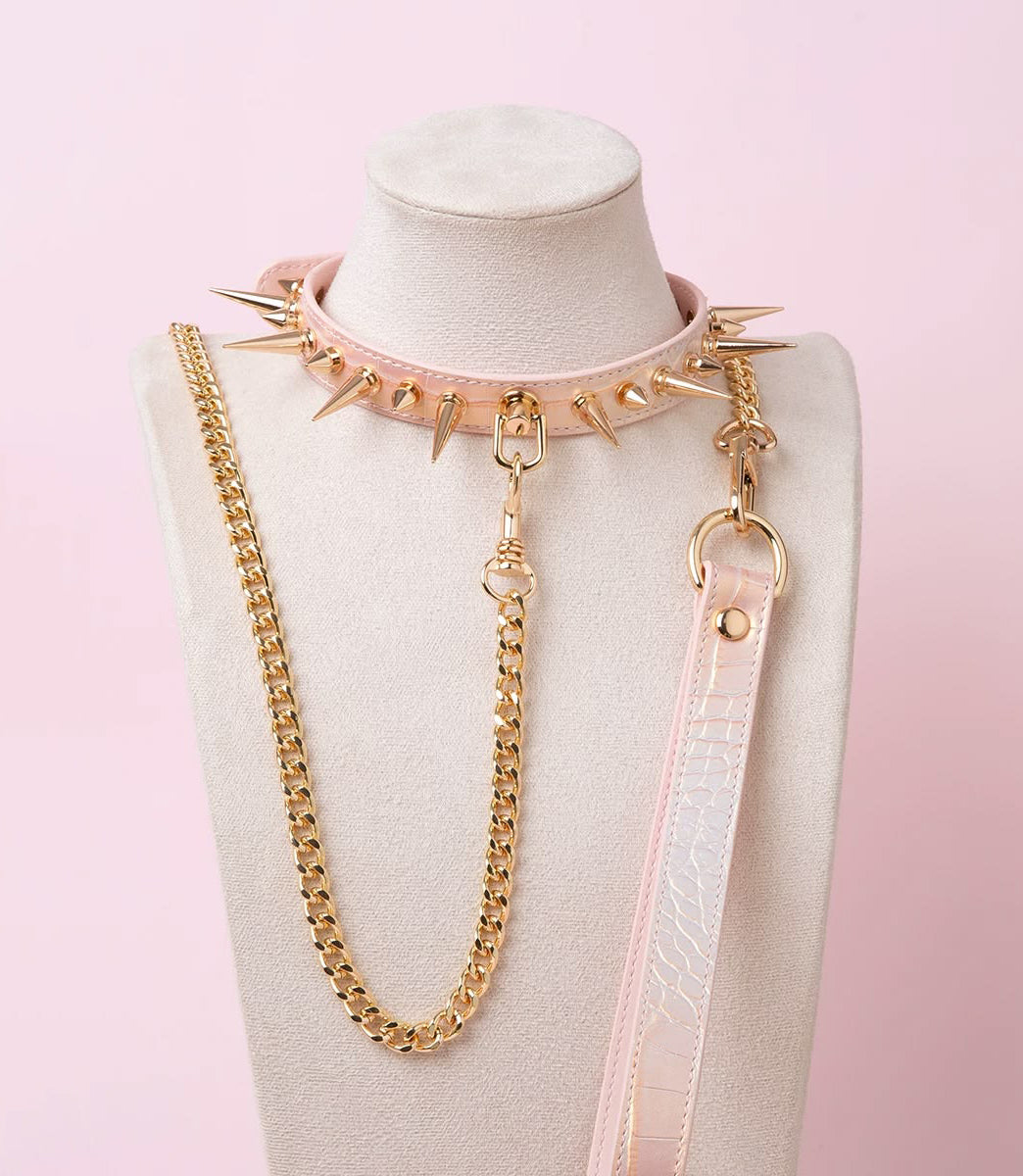 Pink faux crocodile effect leather choker and leash set featuring gold tone alternating length spike studs and central D ring attachment, with detachable chain leash and leather hand strap. Displayed on a white mannequin bust against a pink background for a pastel goth aesthetic.
