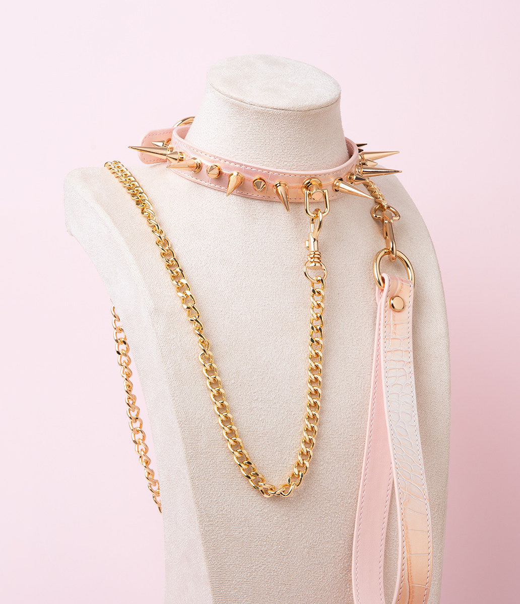 Pink faux crocodile effect leather choker and leash set featuring gold tone alternating length spike studs and central D ring attachment, with detachable chain leash and leather hand strap. Displayed on a white mannequin bust against a pink background for a pastel goth aesthetic.