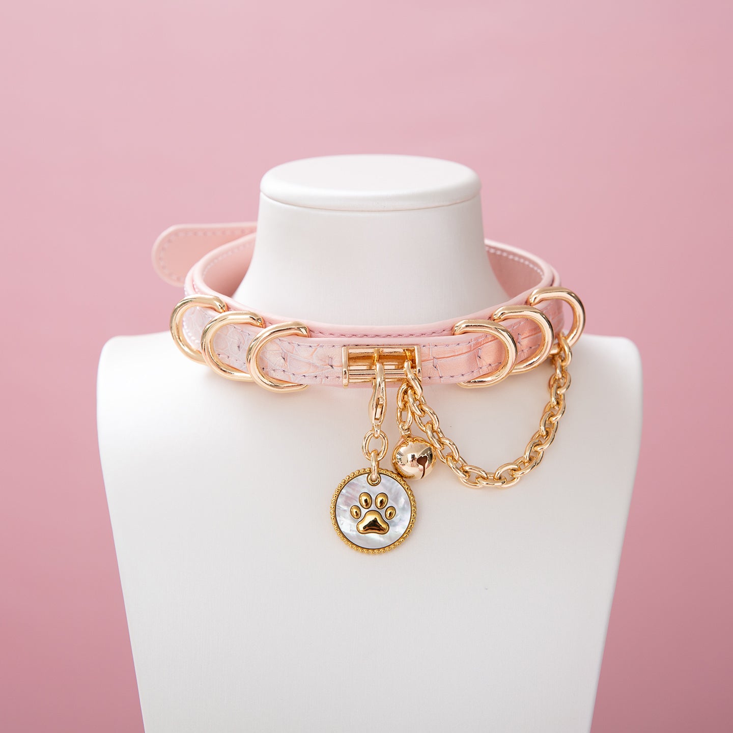 Pink leather choker featuring a gold and white paw print tag, gold tone D rings, chain and bell. Displayed on a white mannequin bust against a pink background for a Harajuku fashion aesthetic.