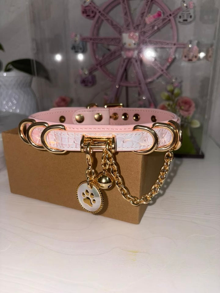 Lifestyle photo of a pink leather choker featuring a white and gold paw print pet tag, gold tone chain, D rings and bell charm. Displayed on a bedroom table for a kawaii aesthetic.
