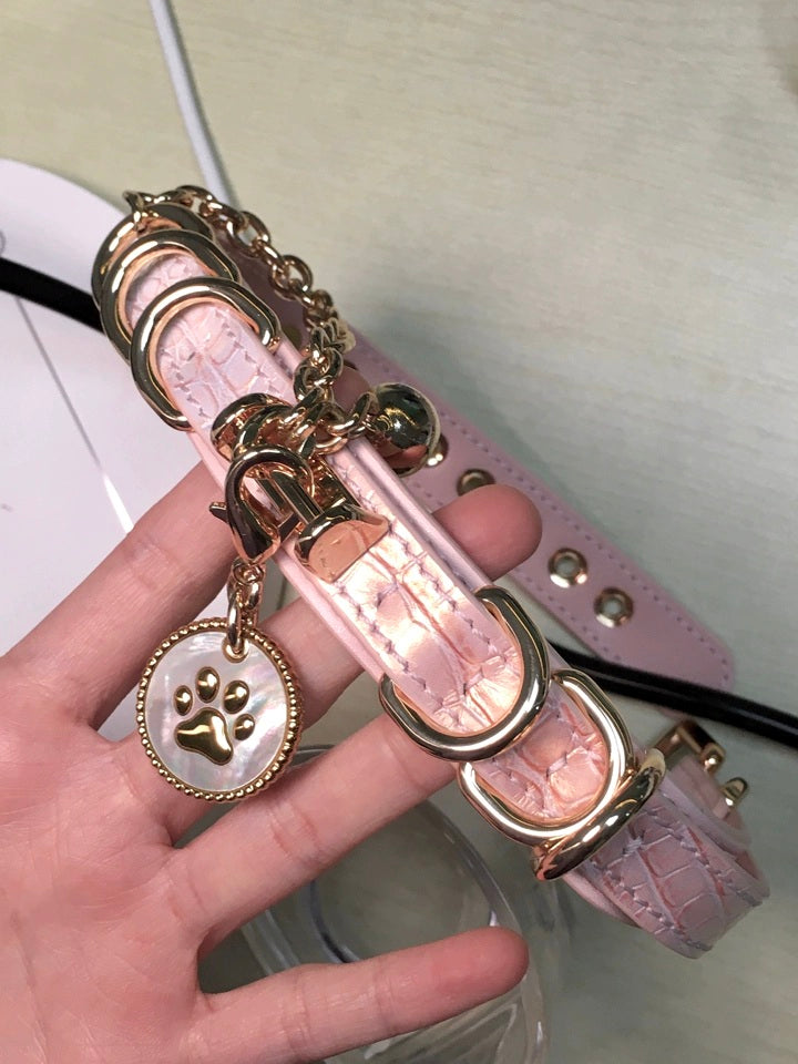 Pink leather choker featuring a white and gold paw print pet tag, gold tone chain, D rings and bell charm. Held by a hand for a casual lifestyle shot.