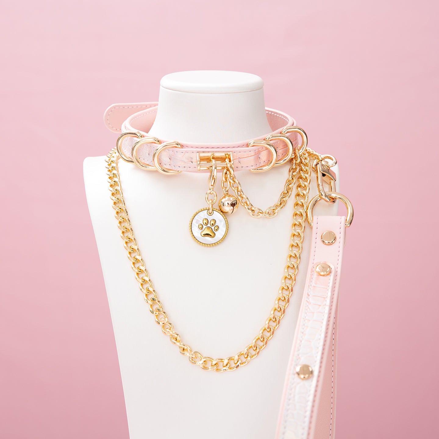 Pink leather choker and leash set featuring a white and gold paw print tag, D rings, chain and bell with detachable chain leash and leather hand strap. Displayed on a white mannequin bust against a pink background for a pastel goth aesthetic.