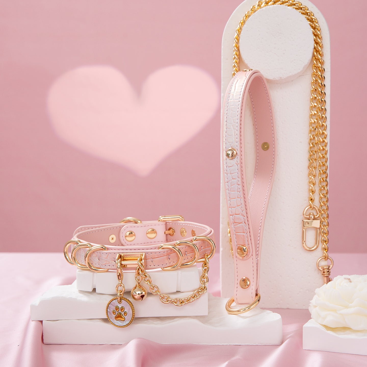 Pink leather choker and leash set featuring a white and gold paw print tag, D rings, chain and bell with detachable chain leash and leather hand strap. Displayed on a white display stand against a pink background for a pastel goth aesthetic.