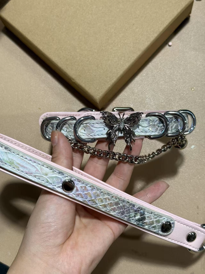 Pink and holographic silver leather choker and leash set featuring a stainless steel plated butterfly charm, D rings, chain and bell with detachable chain leash and leather hand strap. Held by a hand against a table for a casual lifestyle shot.