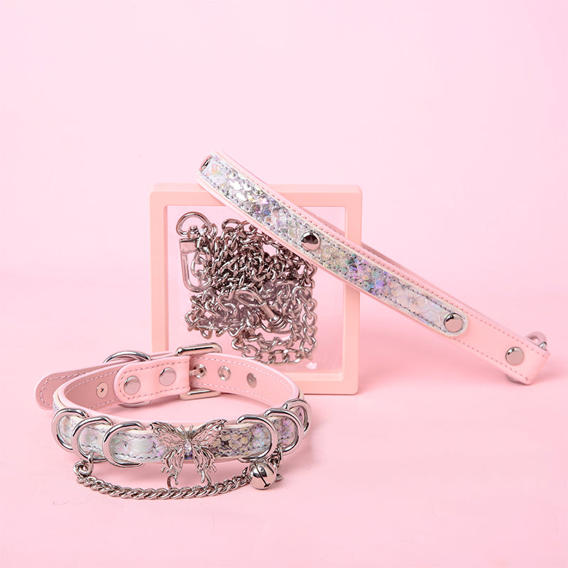 Pink and holographic silver leather choker and leash set featuring stainless steel plated butterfly hardware, D rings, chain and bell with detachable chain leash and leather hand strap. Displayed against a pink background for a pastel goth aesthetic.