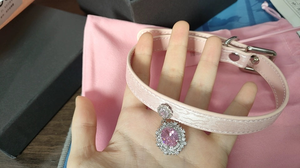 Pink leather choker featuring satin effect leather with a subtle rose print, silver and pink crystal cameo and hardware. Held by a hand against a pink velvet drawstring bag for a casual lifestyle shot.