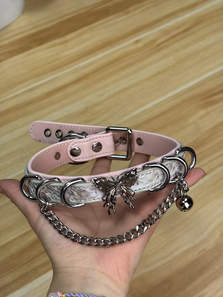 Pink and holographic silver leather choker featuring a stainless steel plated butterfly charm, D rings, chain and bell. Held by a hand against a wooden table for a casual lifestyle aesthetic.