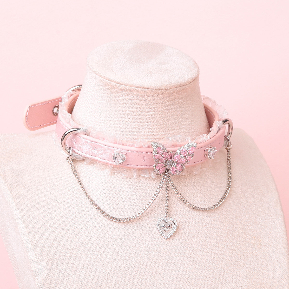 Pink leather choker featuring white ruffled lace trim, diamante butterfly and heart studs, silver tone D rings and chain. Displayed on a white mannequin bust against a pink background for a kawaii goth aesthetic.