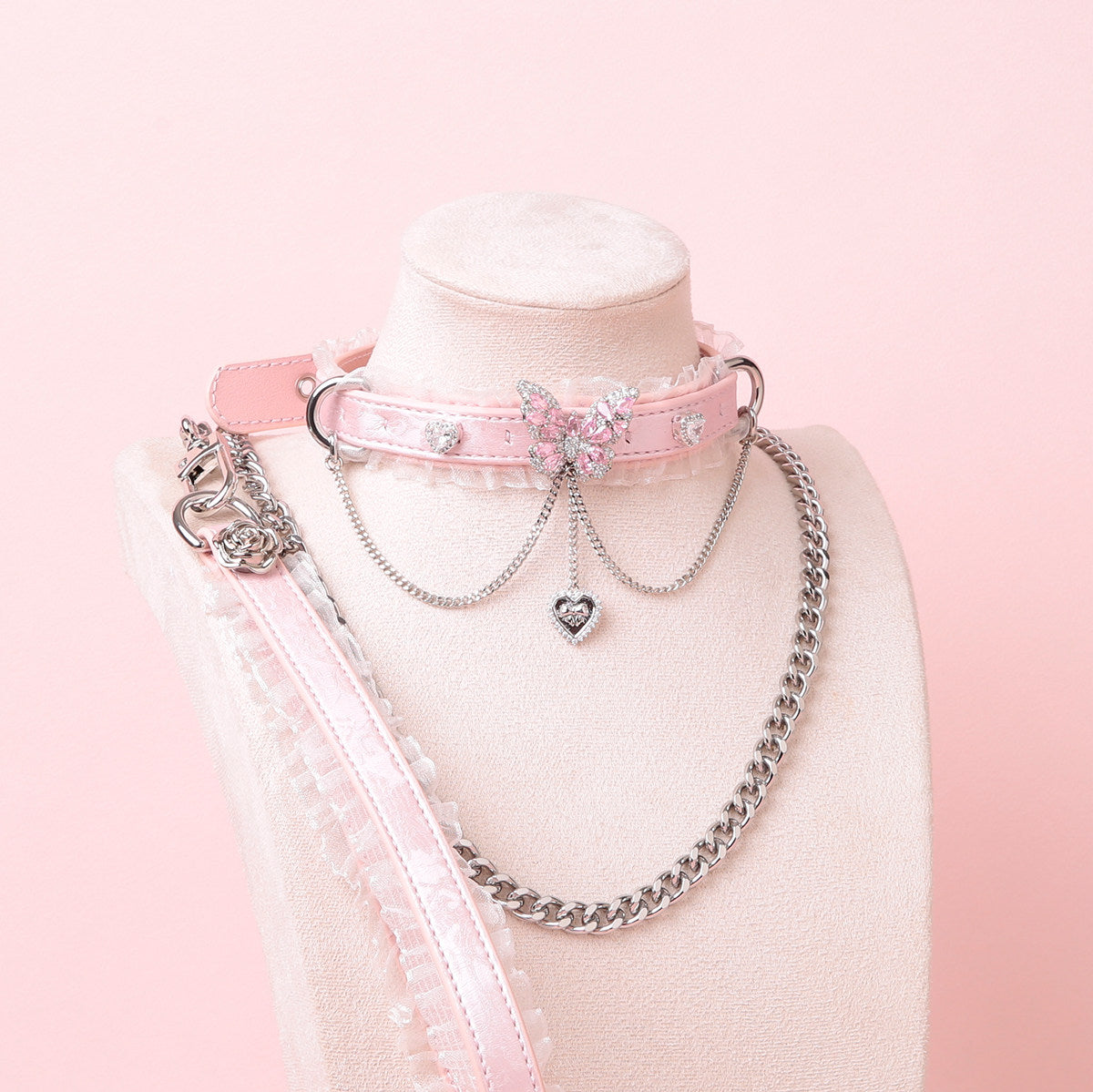 Pink leather choker featuring white ruffled lace trim, diamante butterfly and heart charms, silver tone chain and D rings with detachable silver chain leash and pink and white leather hand strap. Displayed on a white mannequin bust against a pink background for a pastel goth aesthetic.