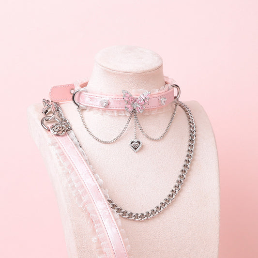 Pink leather choker featuring white ruffled lace trim, diamante butterfly and heart charms, silver tone chain and D rings with detachable silver chain leash and pink and white leather hand strap. Displayed on a white mannequin bust against a pink background for a pastel goth aesthetic.