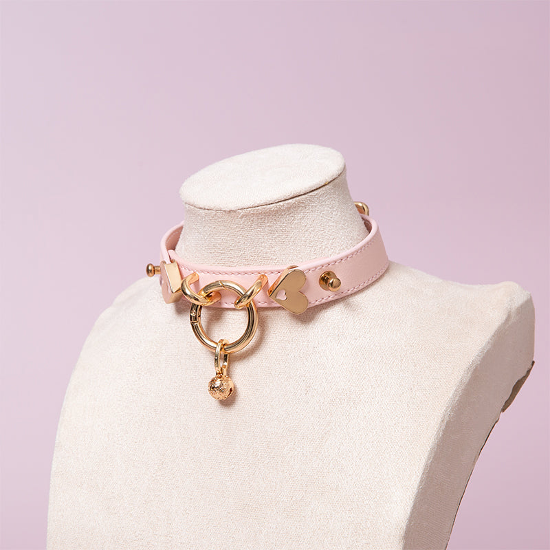 Pink leather choker featuring gold tone O ring, heart hardware and detachable bell charm. Displayed on a white mannequin bust against a light pink background for a pastel goth aesthetic.