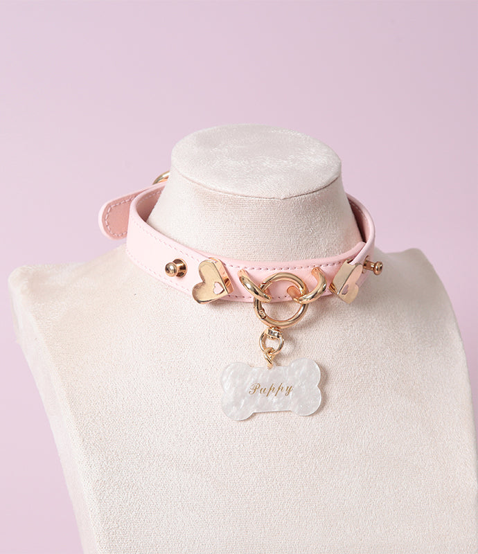 Pink leather choker featuring gold tone O ring, heart hardware and detachable white acrylic bone shaped pet tag, with custom engraving available. Displayed on a white mannequin bust against a light pink background for a pastel goth aesthetic.