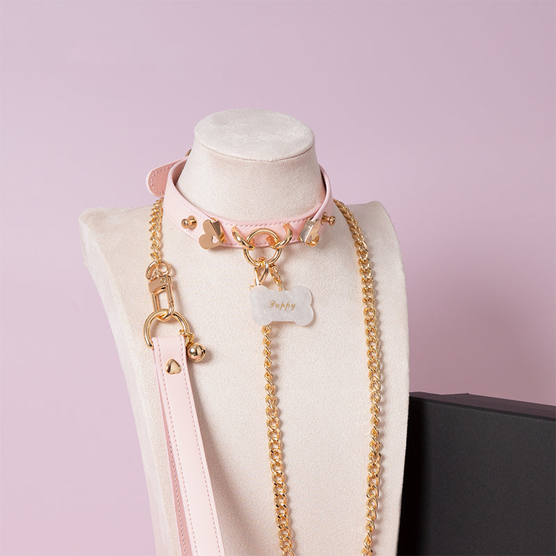 Pink leather choker featuring gold tone hardware, custom engraved bone shaped pet tag, heavy duty chain leash and leather hand strap. Displayed on a white mannequin bust against a light pink background for a pastel goth aesthetic.