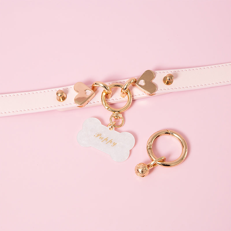 Close-up photo of a pink leather choker featuring a light gold O ring, heart hardware and two detachable bell and bone shaped pet tag charms. Displayed on a light pink background for a kawaii aesthetic.