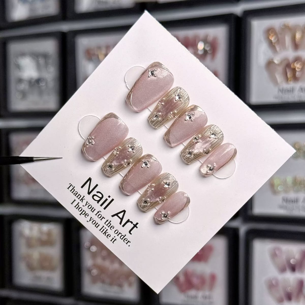 A set of 10 press-on nails displayed on a white card, featuring a combination of pale pink nude and glittery gold designs with small rhinestone embellishments. The card is held by tweezers, with a blurred background of other framed nail art samples.