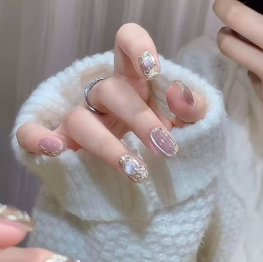 A close-up of manicured fingernails on a hand wearing a white fuzzy knit sweater. The nails are glossy nude pink cat eye with glittery gold patterns and iridescent pearl-like accents.