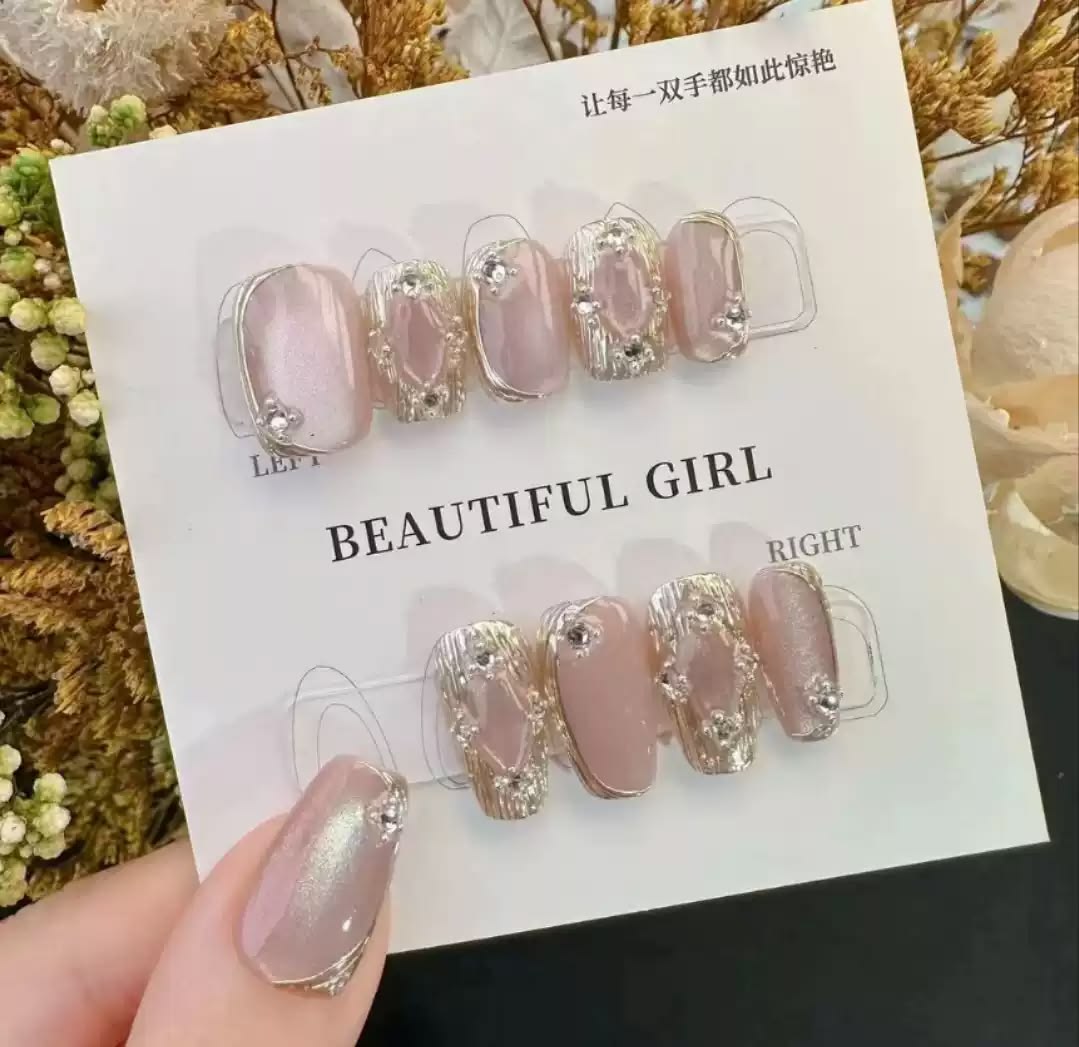 A set of 10 press-on nails displayed on a white card, featuring a combination of pale pink nude and glittery gold designs with small rhinestone embellishments. The card is held by a hand, with a blurred floral background.