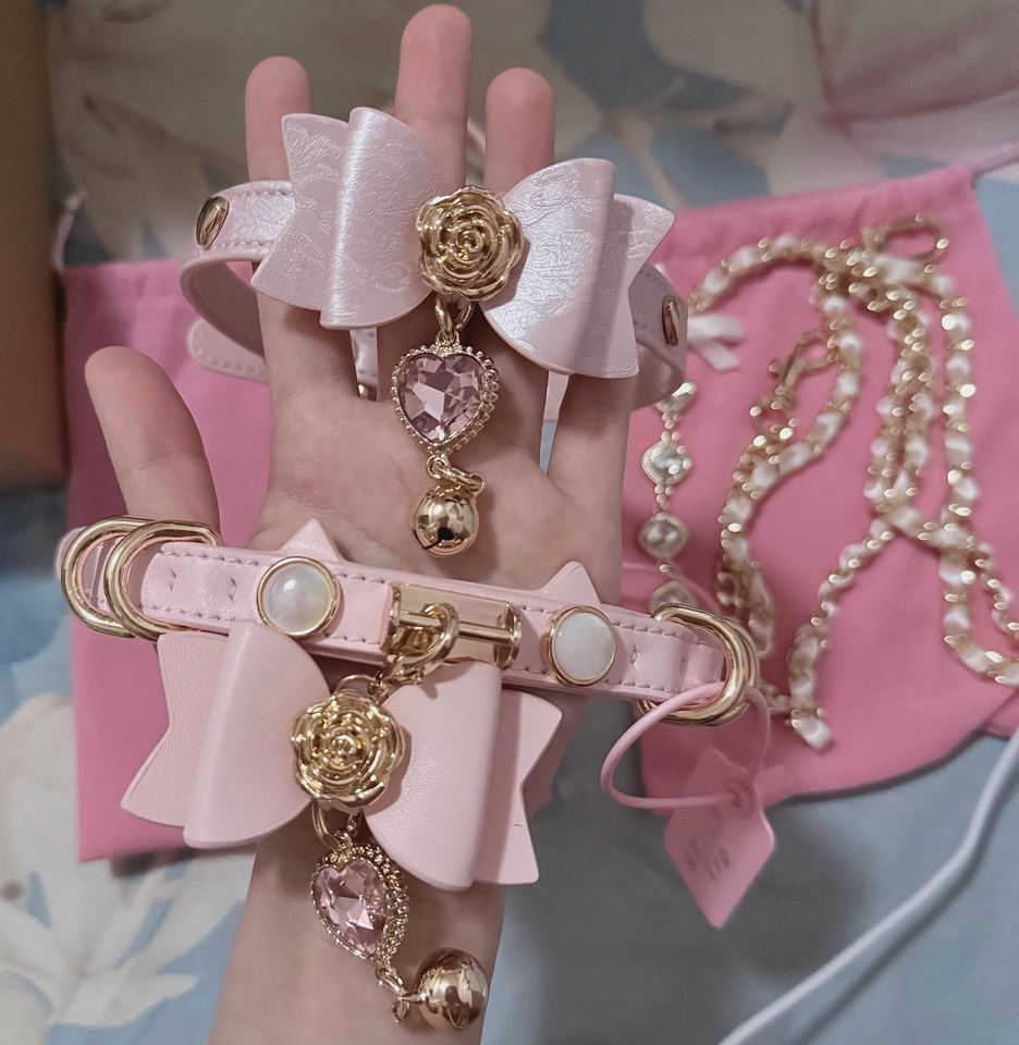 Two pink leather chokers featuring ribbon bow and cat ear shaped straps, gold tone hardware, faux mother of pearl studs and detachable pink and gold crystal heart and bell pendants. Held by a hand against a floral background showing the matching gold and diamante chain leashes for a kawaii BDSM aesthetic.