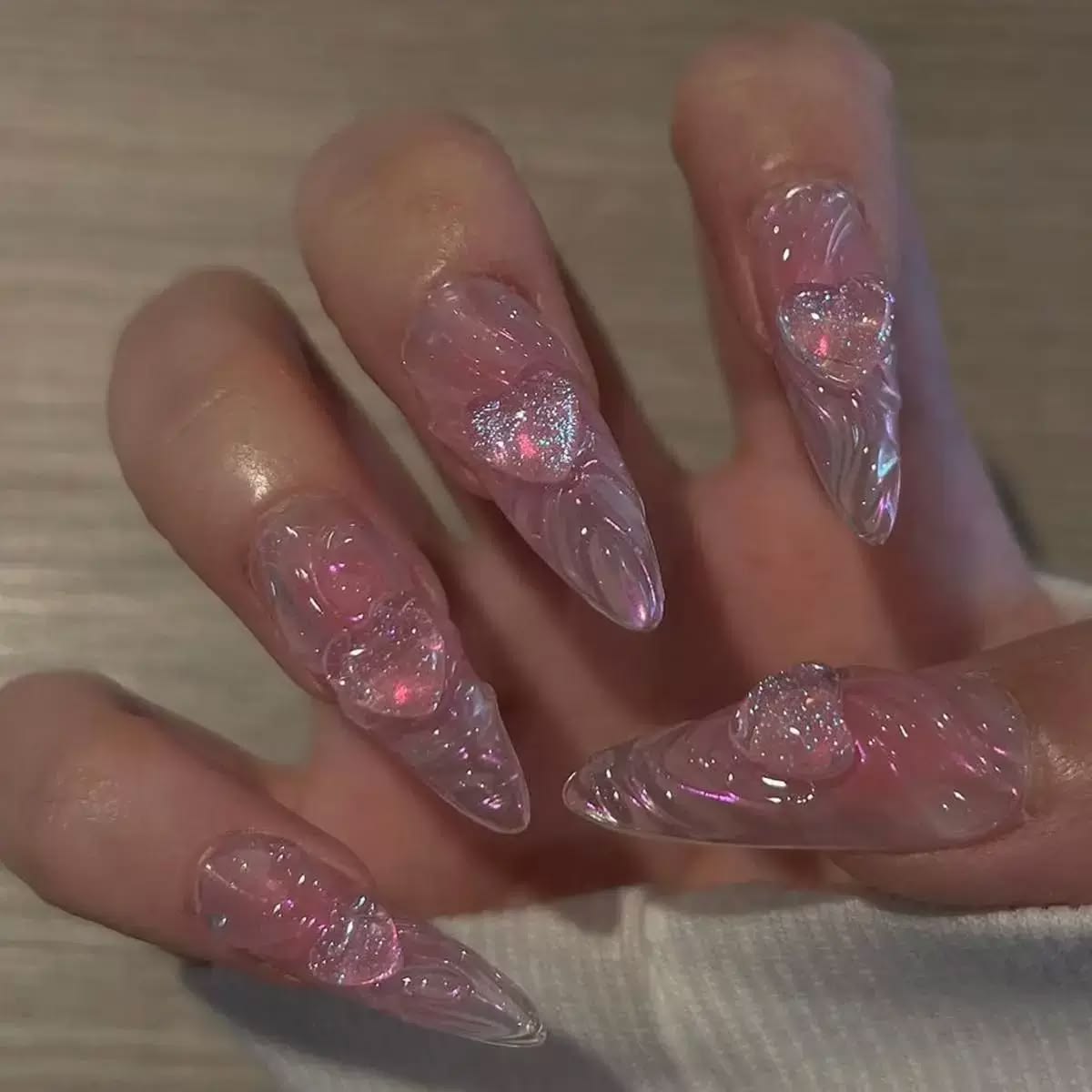 A close-up of a hand with long, sculpted stiletto false nails decorated with pink and clear 3D swirls and glittery pink hearts.
