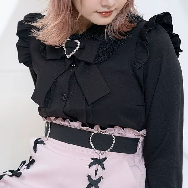 Model photo of a pink high waisted skirt featuring a ruffled waist with side zip fastening, diamante heart shaped buckle belt, black satin ribbon corset lacing and pleated a-line design, styled with a black ruffled blouse for a Jirai Kei fashion aesthetic.
