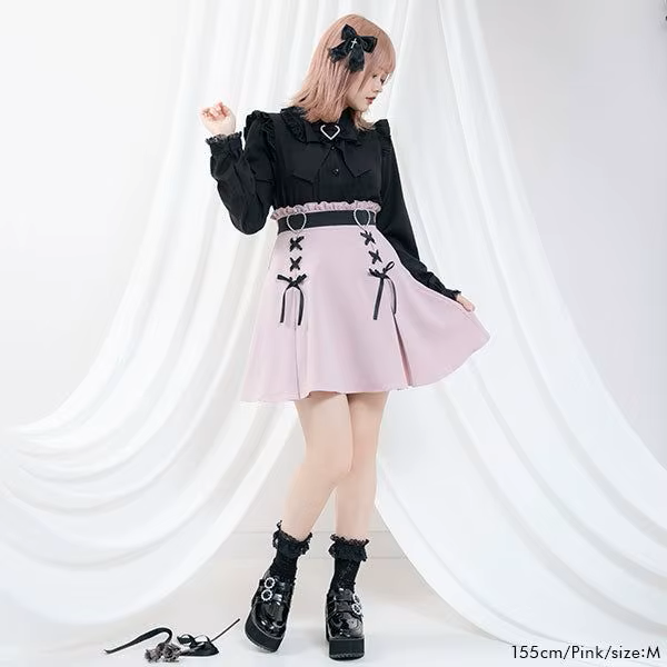 Modelled photo of a pink high waisted skirt featuring a ruffled waist, diamante heart shaped buckle belt, black satin ribbon corset lacing and pleated a-line design, styled with a black blouse and chunky black Mary Jane shoes for a Jirai Kei fashion aesthetic.