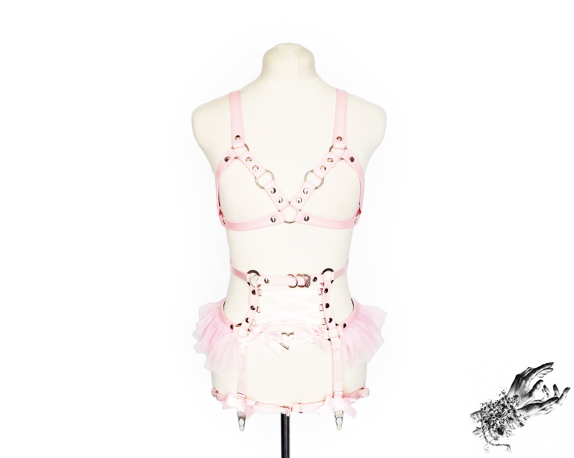 Pink vegan leather harness set featuring silver O ring hardware, pink ruffled tulle accents at the waist and corset lacing, displayed on a mannequin against a white background.