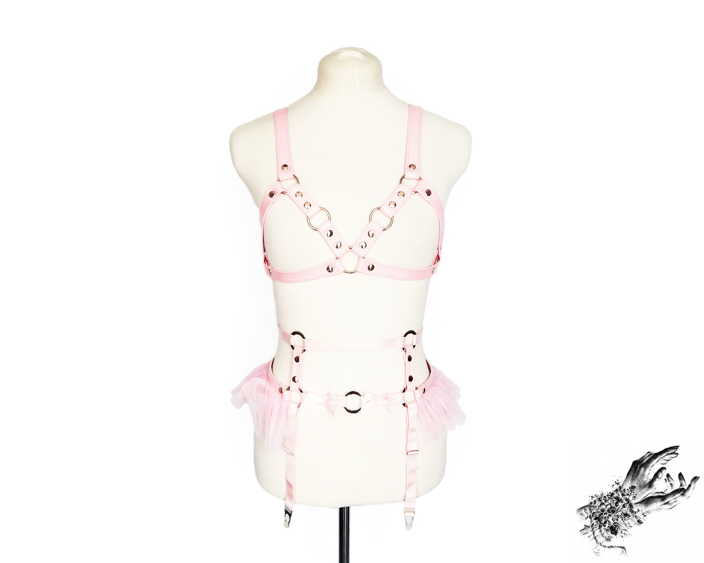 Pink faux leather harness set featuring a separate harness bra and garter belt with silver O ring hardware, ruffled tulle and bow appliques, displayed on a mannequin against a white background.