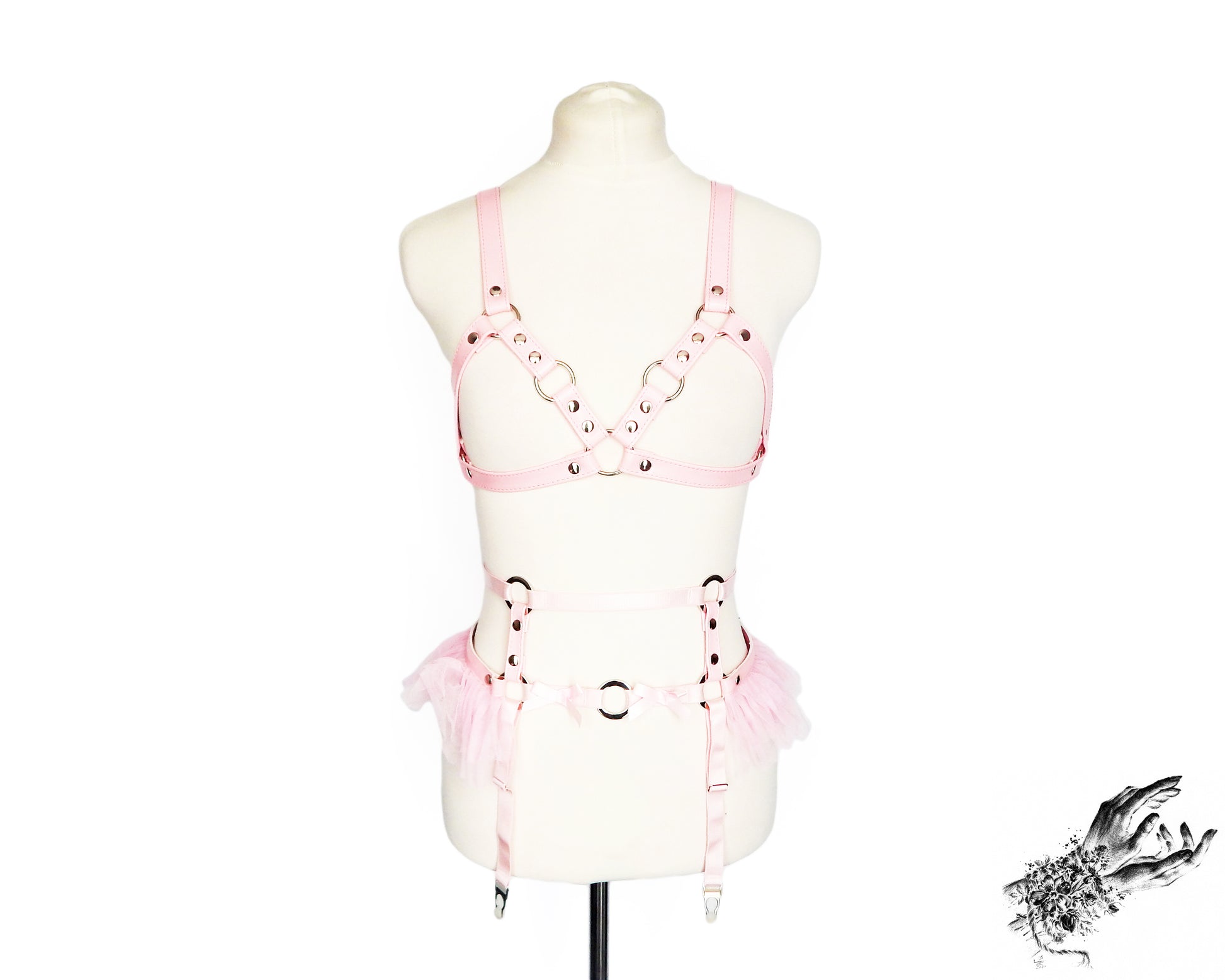 Pink faux leather harness set featuring a separate harness bra and garter belt with silver O ring hardware, ruffled tulle and bow appliques, displayed on a mannequin against a white background.