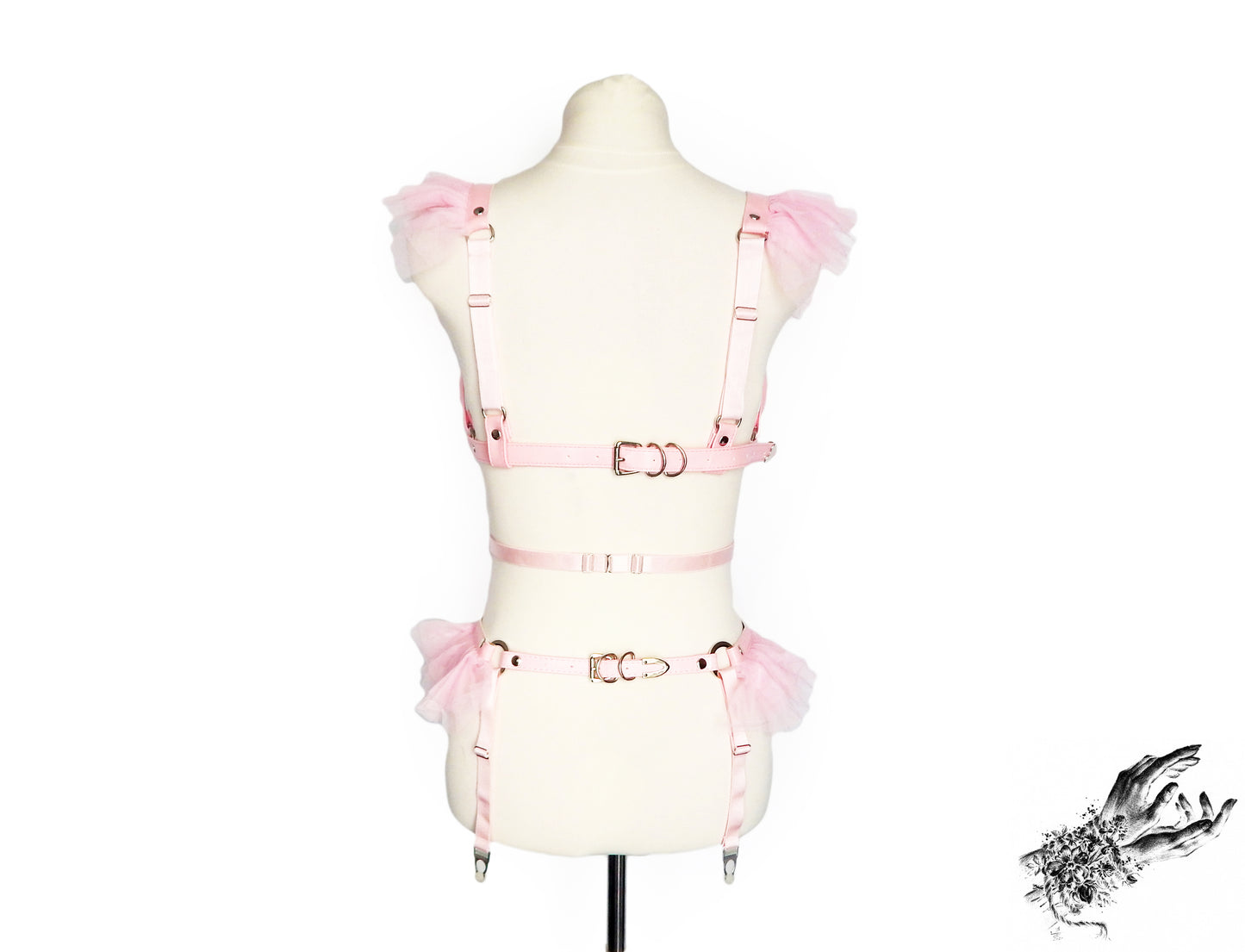 A light pink vegan leather harness set with matching pink tulle ruffle accents at the shoulders and hips and adjustable elastic and buckle straps, displayed on a cream mannequin torso against a white background.