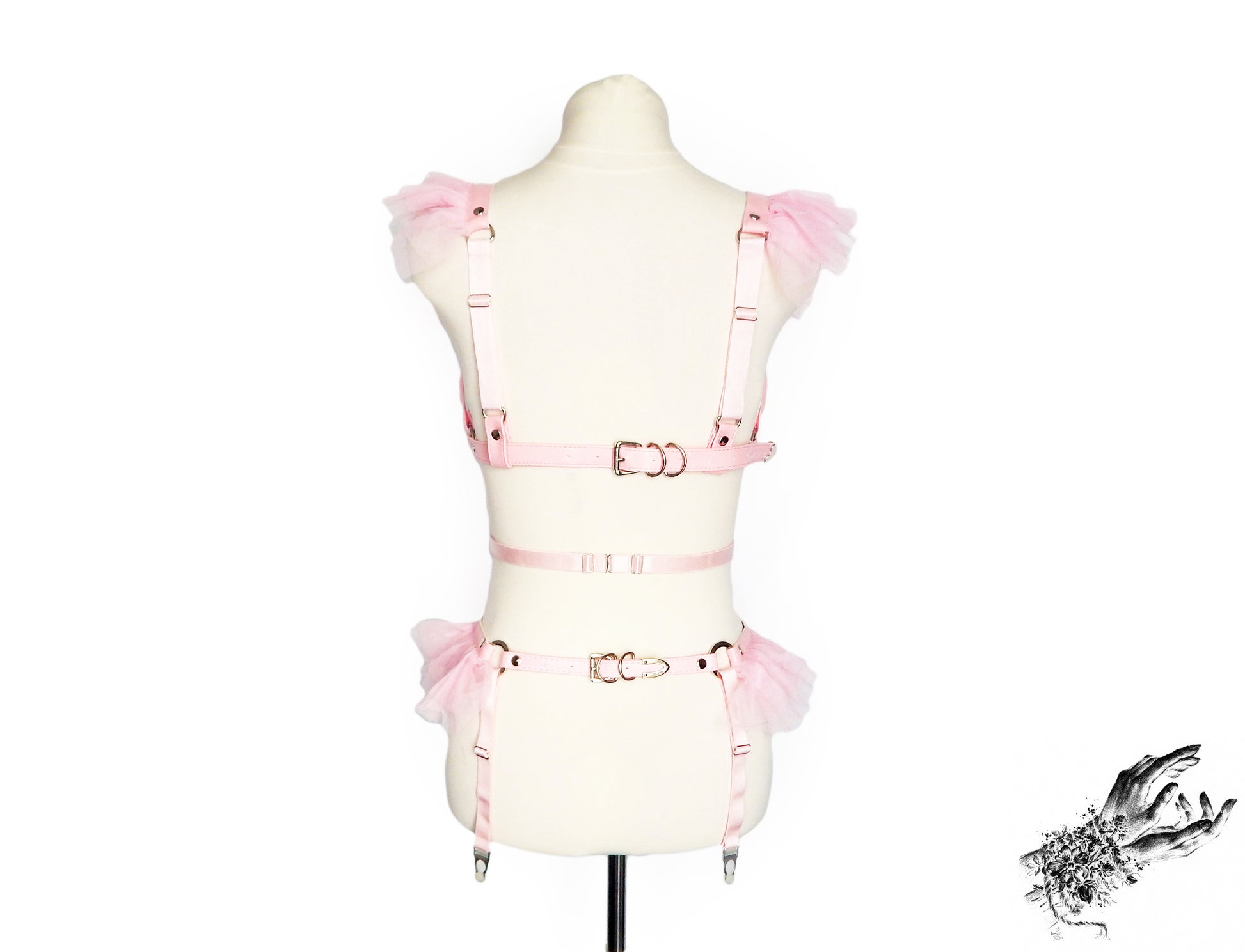A light pink vegan leather harness set with matching pink tulle ruffle accents at the shoulders and hips and adjustable elastic and buckle straps, displayed on a cream mannequin torso against a white background.