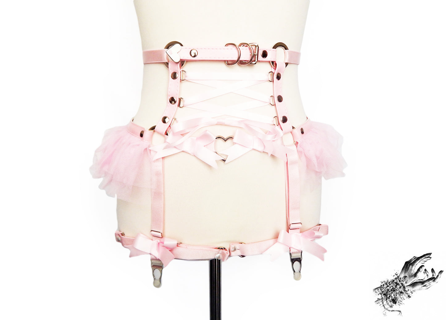 Pink faux leather garter belt featuring silver O ring hardware, pink ruffled tulle accents at the hips and corset lacing, displayed on a mannequin against a white background.