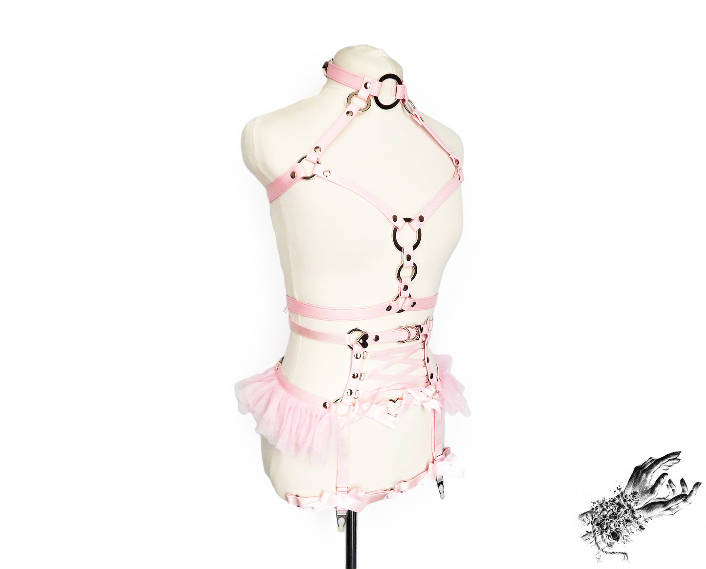 Pink faux leather harness set featuring a separate chest harness and garter belt with silver O ring hardware, pink corset lacing and ruffed tulle accents at the waist, displayed on a mannequin against a white background.