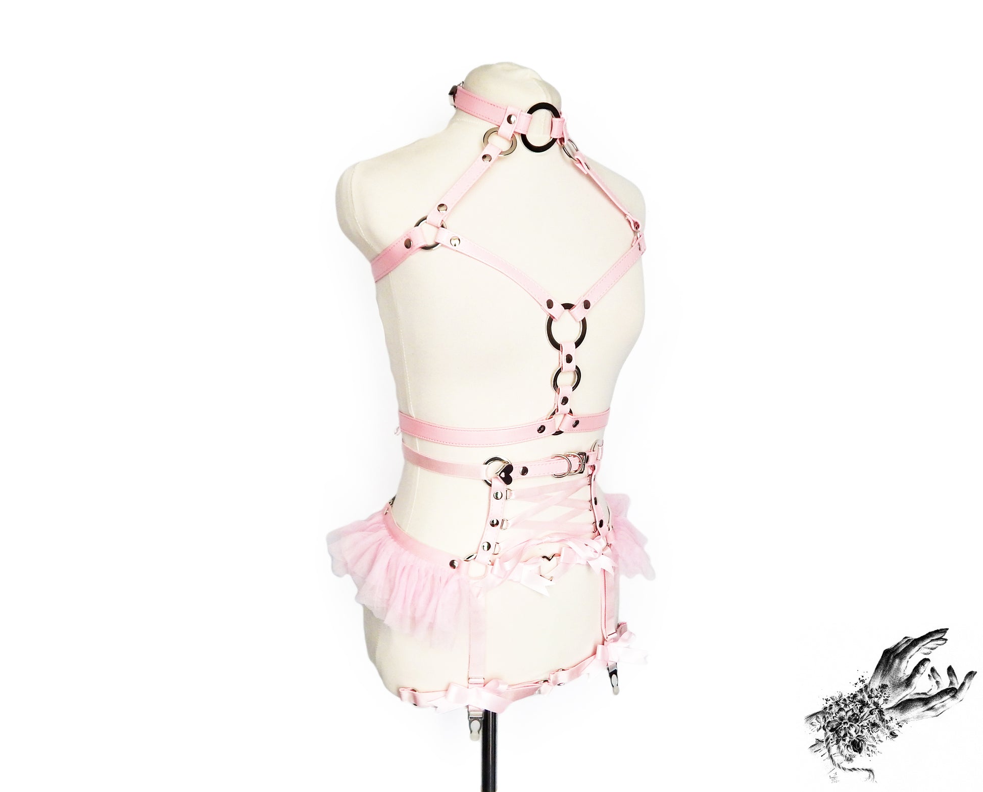 Pink faux leather harness set featuring a separate chest harness and garter belt with silver O ring hardware, pink corset lacing and ruffed tulle accents at the waist, displayed on a mannequin against a white background.