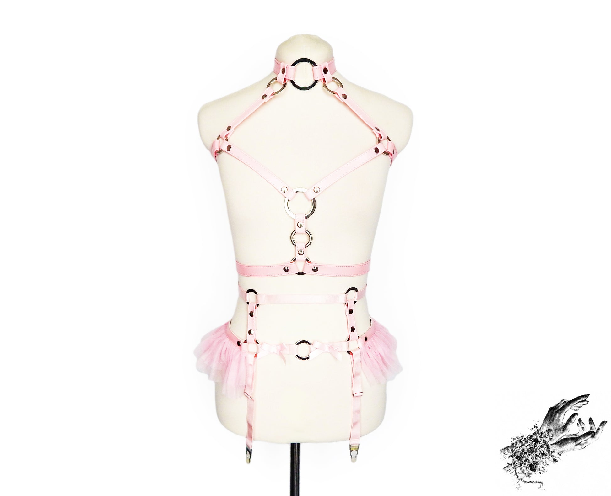 Front view of a pink vegan leather harness set featuring a separate top harness and garter belt with silver O ring hardware, pink ruffled tulle accents at the waist and adjustable straps, displayed on a mannequin against a white background.